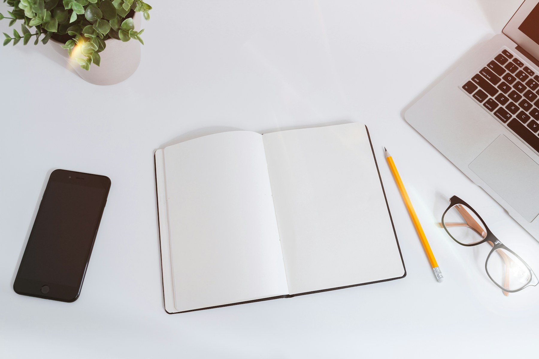 An open notebook, laptop, phone, pencil and glasses arranged on a bright desk