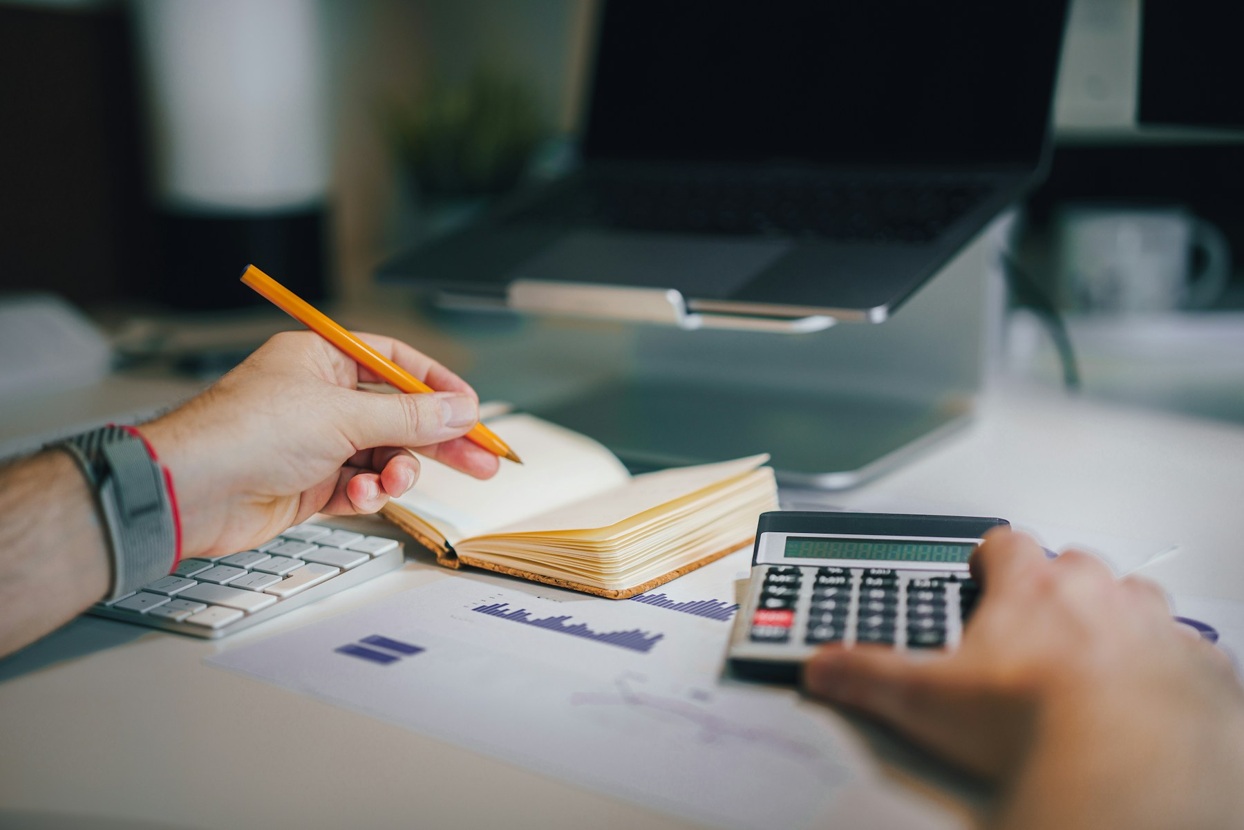 Hands using a calculator beside a cream notebook and printed charts